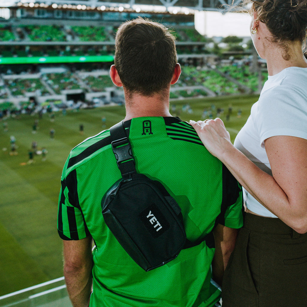 A man in a bright green and black sports jersey seen from behind, wearing a black YETI crossbody bag across his chest, standing in a stadium with a woman beside him.