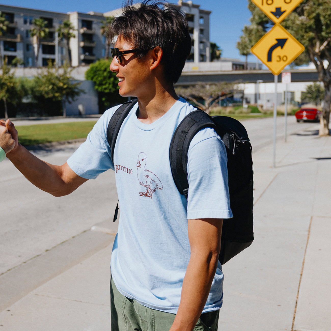 Man wearing a light blue t-shirt with a graphic design and sunglasses, standing on a sidewalk with a cityscape background.