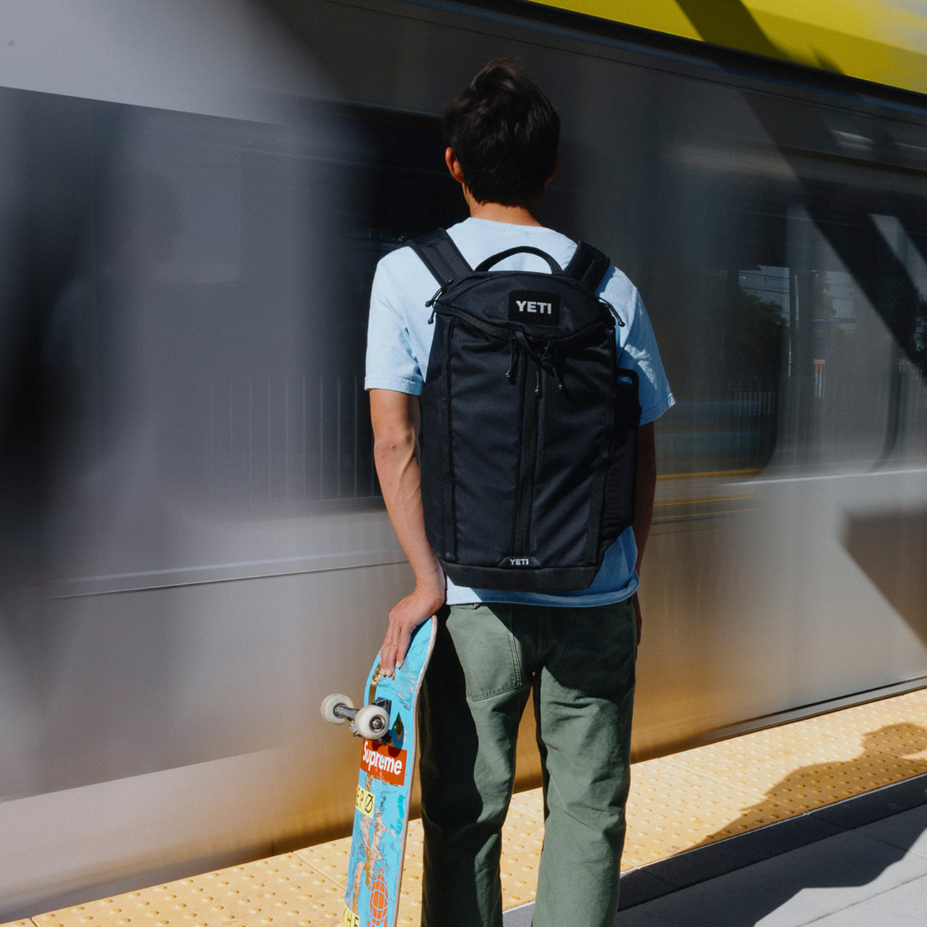 Person with a black YETI backpack holding a skateboard at a train station.