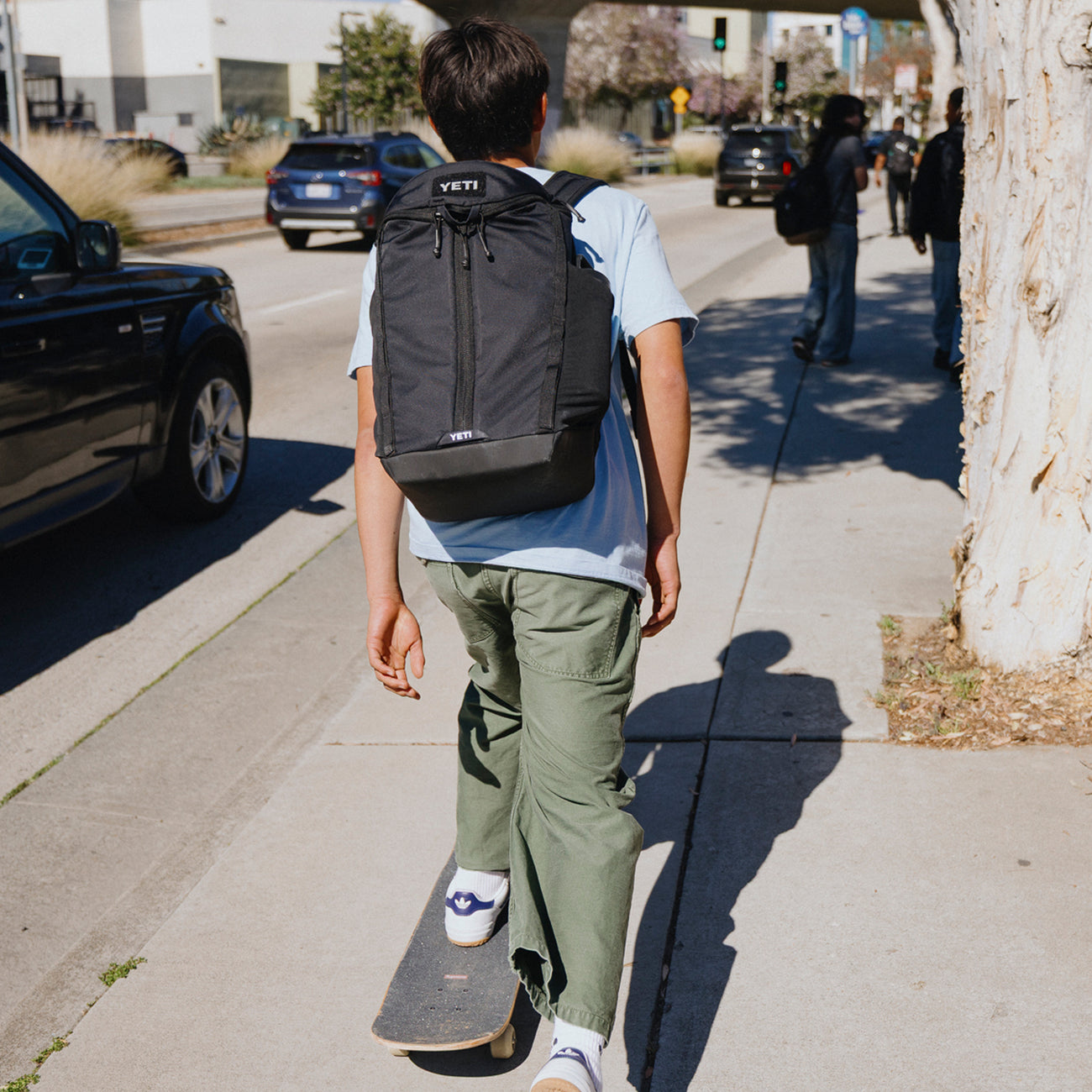 Person walking with a skateboard on a city street, wearing a black backpack.